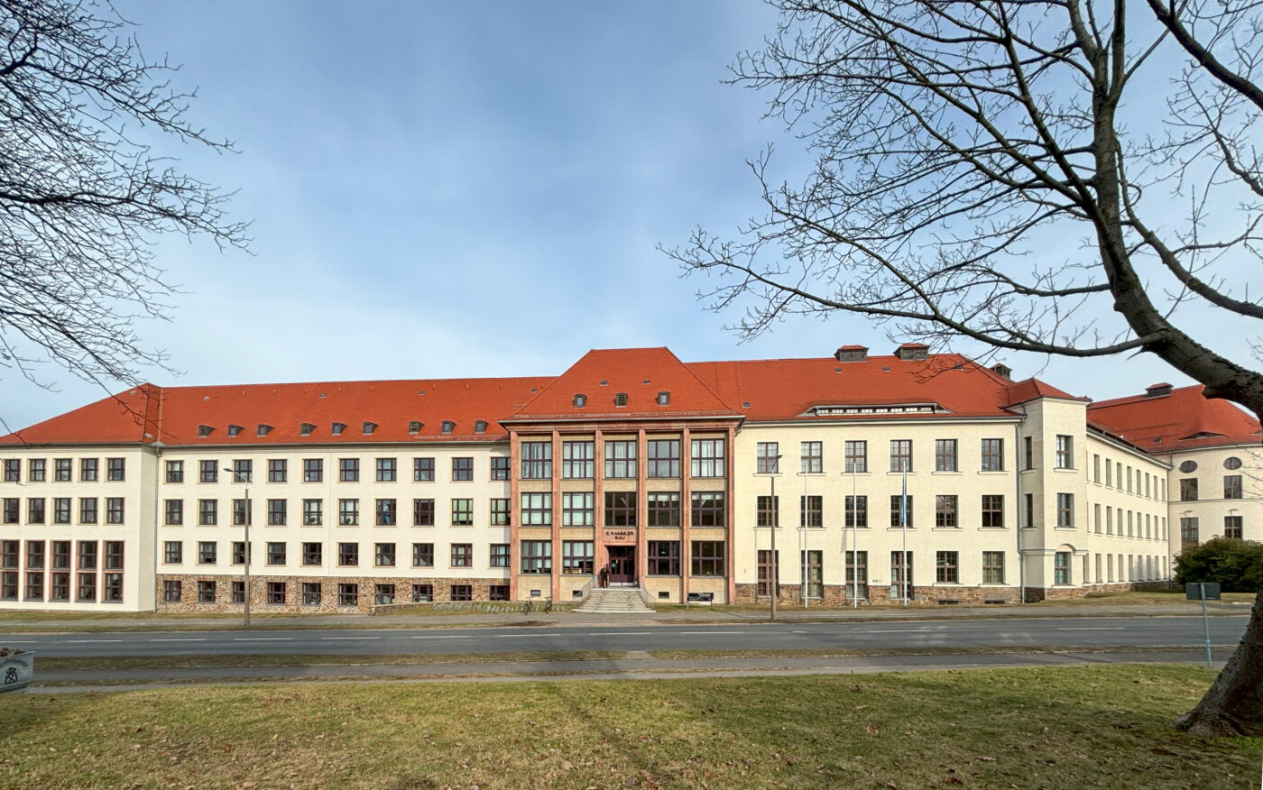 Die Hauptfassade des Erich-Rammler-Baus. zum Haupteingang führt eine Treppe, die Fassade ist hell und große Fenster in Achsen und das Dach ist rot.