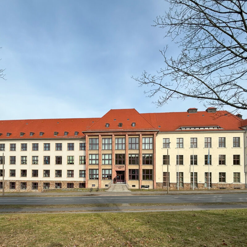 Die Hauptfassade des Erich-Rammler-Baus. zum Haupteingang führt eine Treppe, die Fassade ist hell und große Fenster in Achsen und das Dach ist rot.